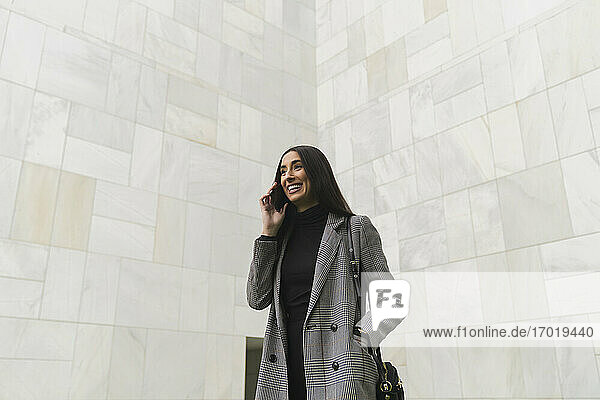 Lächelnde Geschäftsfrau mit Händen in den Taschen  die mit einem Mobiltelefon an der Wand spricht