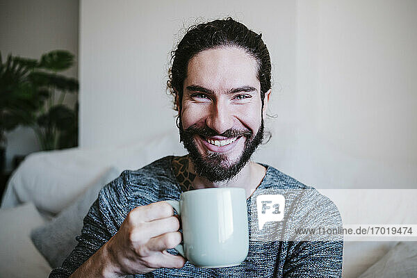 Junger Mann lächelnd beim Kaffeetrinken zu Hause sitzend