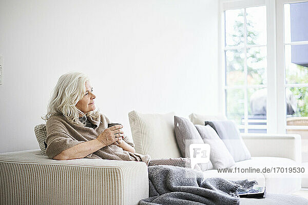 Nachdenkliche Frau mit langen weißen Haaren hält eine Kaffeetasse  während sie auf dem Sofa in einer Wohnung sitzt