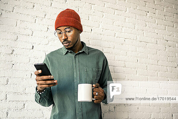 Mann mit Kaffeetasse und Smartphone vor weißer Backsteinwand