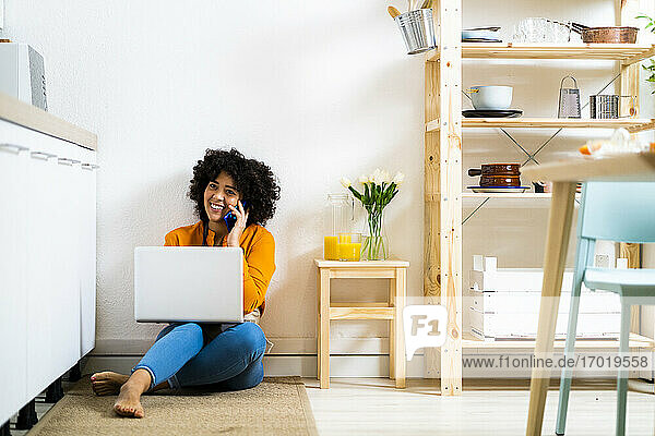 Lächelnde junge Frau mit Laptop  die mit ihrem Handy telefoniert  während sie zu Hause auf dem Boden sitzt
