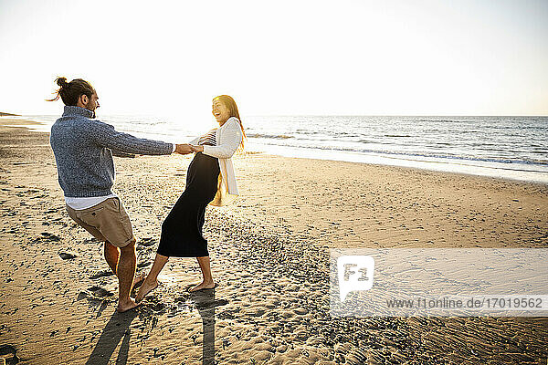 Cheerful couple holding hands while dancing at beach against clear sky during sunny day