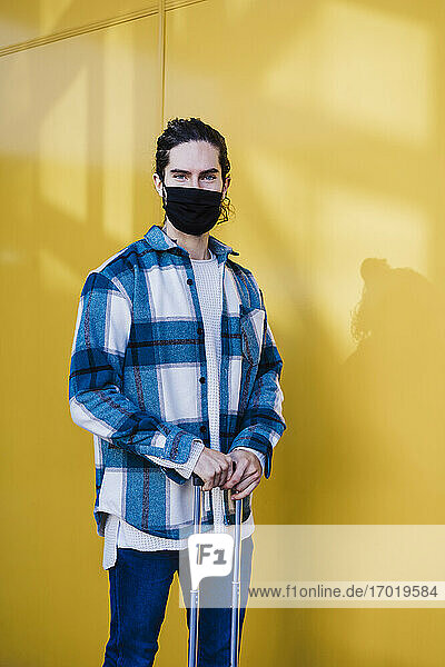 Junger Mann mit Gesichtsschutzmaske steht mit Gepäck vor einer gelben Wand