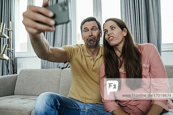 Happy couple making faces while taking selfie while sitting on sofa at home