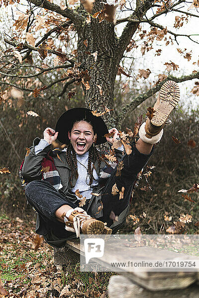 Smiling teenage girl in hat sitting on plank in Autumn landscape