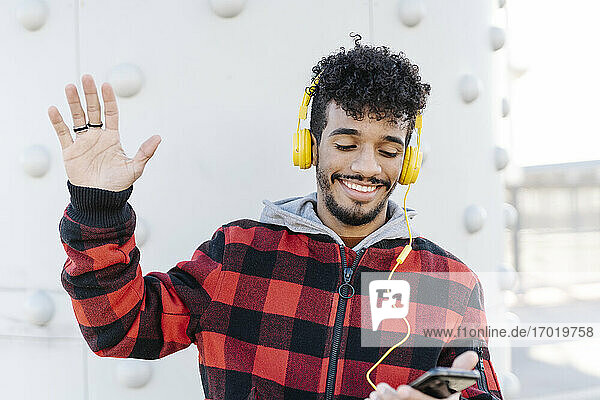 Lächelnder Mann mit Kopfhörern  der mit der Hand winkt  während er ein Mobiltelefon benutzt und an der Wand steht