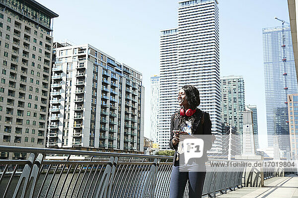 Geschäftsfrau mit Kopfhörern und Mobiltelefon schaut weg  während sie auf einer Brücke in der Stadt spazieren geht