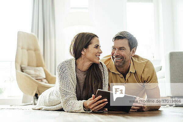 Smiling man and woman with digital tablet looking at each other while lying in home