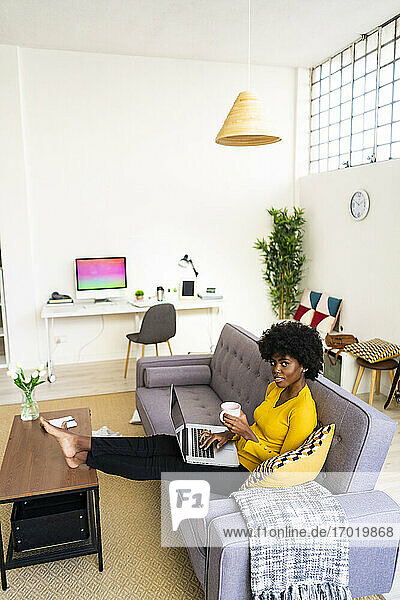 Junge Frau mit Laptop trinkt Kaffee  während sie zu Hause auf dem Sofa sitzt