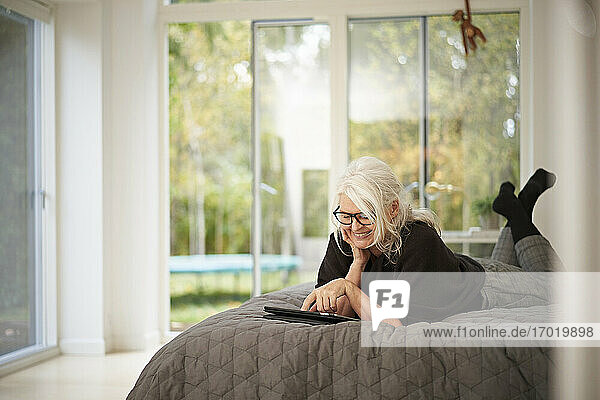 Lächelnde ältere Frau  die ein digitales Tablet benutzt  während sie zu Hause auf dem Bett liegt