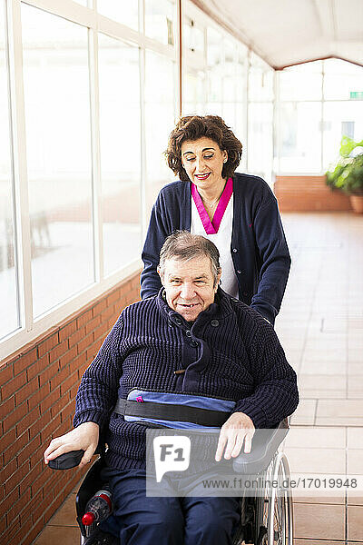 Ältere Krankenschwester hilft behindertem Mann im Rollstuhl sitzend auf dem Flur eines Rehabilitationszentrums