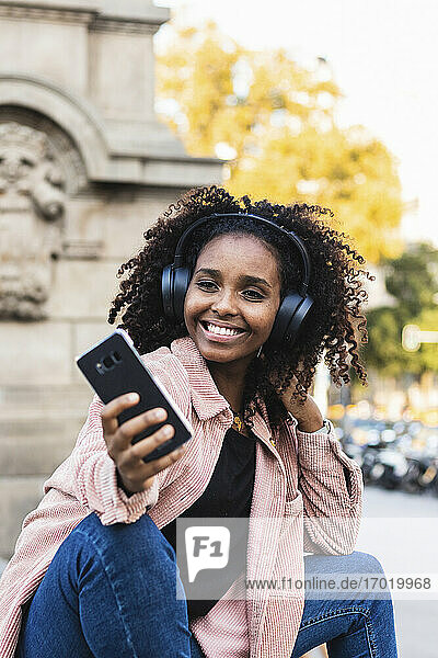 Smiling fashionable young woman taking selfie while listening music in city