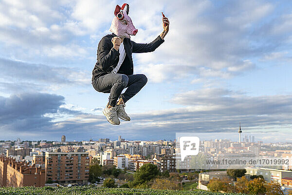 Male professional wearing pig mask taking selfie from smart phone while jumping in city against sky