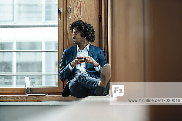Junger Geschäftsmann hält Kaffeetasse und schaut weg  während er im Büro am Fenster sitzt