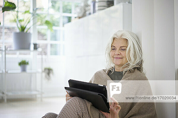 Smiling woman with white hair using digital tablet while sitting in living room at apartment