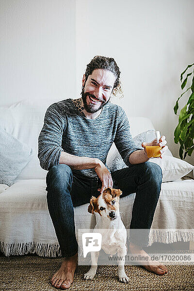 Lächelnder junger Mann hält ein Saftglas in der Hand und sitzt mit seinem Hund auf dem Sofa zu Hause