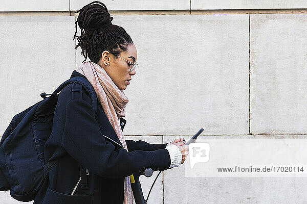 Jamaikanische Frau mit Dreadlocks benutzt ihr Smartphone  während sie sich auf einen Roller an der Wand lehnt
