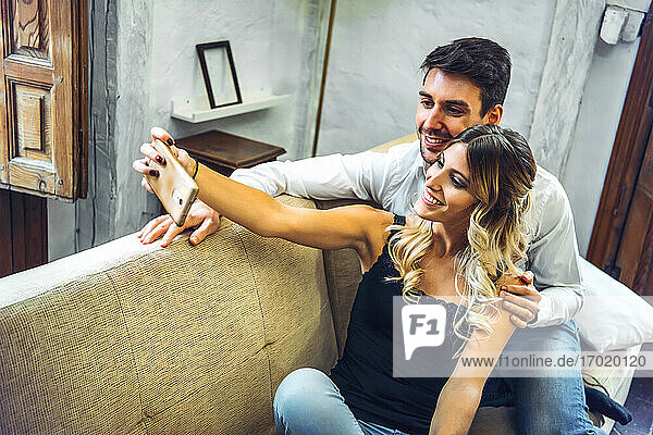 Lächelndes junges Paar nimmt Selfie auf Couch im Wohnzimmer zu Hause