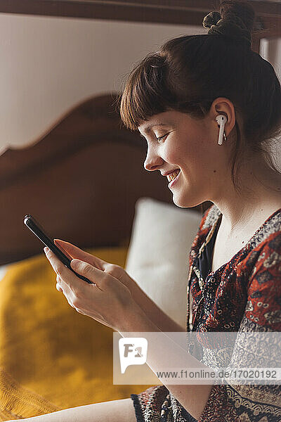 Lächelnde Frau  die einen Videoanruf auf einem Smartphone tätigt  während sie im Schlafzimmer zu Hause sitzt