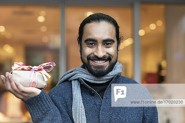 Nahaufnahme eines lächelnden jungen Mannes  der ein Weihnachtsgeschenk vor einem Gebäude zeigt