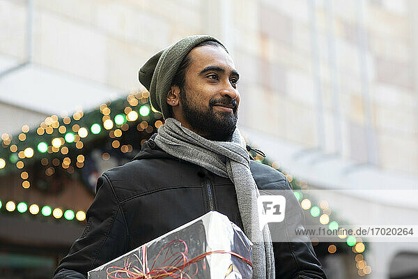 Lächelnder junger Mann mit Weihnachtsgeschenk in der Hand  während er an einem Gebäude in der Stadt steht
