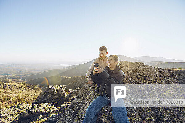 Friends using smart phone while sitting on mountain against clear sky