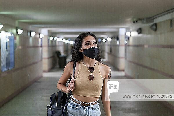 Junge Frau mit Gesichtsmaske  die eine Tasche trägt  während sie in einer unterirdischen Bahnhofspassage geht