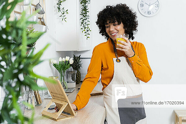 Lächelnde Frau mit Schürze  die Orangensaft trinkt  während sie ein digitales Tablet zu Hause benutzt