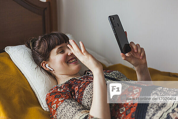 Glückliche Frau  die einen Videoanruf über ihr Smartphone tätigt  während sie zu Hause im Schlafzimmer liegt