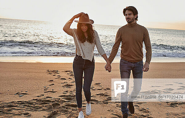 Young woman wearing hat holding hand of man while walking against sea