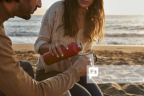 Frau gießt Kaffee in eine Tasse und sitzt neben einem Mann am Strand