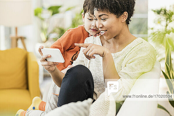 Lächelnde Mutter zeigt ihrem Sohn das Handy  während sie zu Hause sitzt