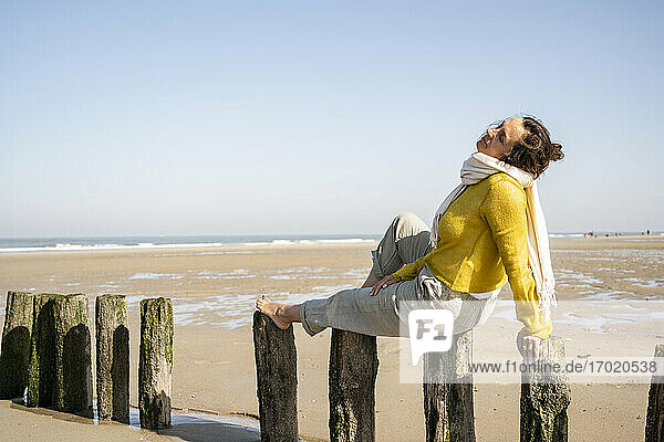 Reife Frau mit geschlossenen Augen sitzt auf Holzpfosten am Strand gegen klaren Himmel