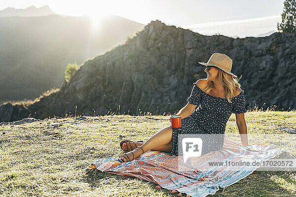 Smiling woman with coffee cup day dreaming while sitting on picnic blanket on sunny day