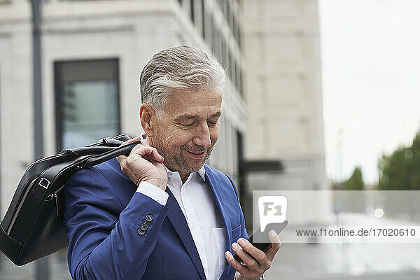 Senior male entrepreneur holding laptop bag using smart phone in city