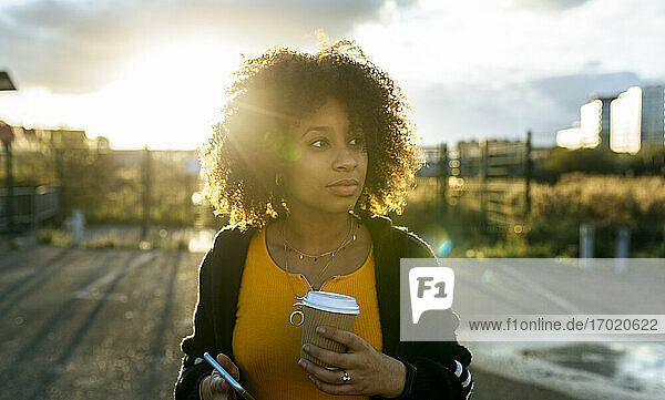 Young woman with afro hair holding coffee and smart phone looking away against sky