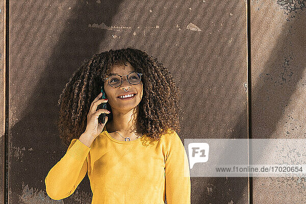 Lächelnde junge Frau  die an einem sonnigen Tag mit ihrem Handy an der Wand telefoniert