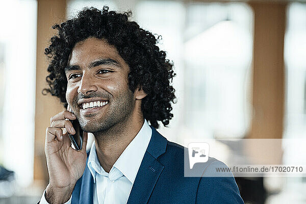 Fröhlicher männlicher Unternehmer  der im Büro mit dem Handy telefoniert und dabei wegschaut