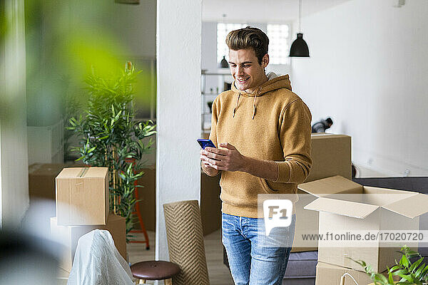 Lächelnder junger Mann  der in einem unordentlichen Wohnzimmer während eines Umzugs ein Mobiltelefon benutzt