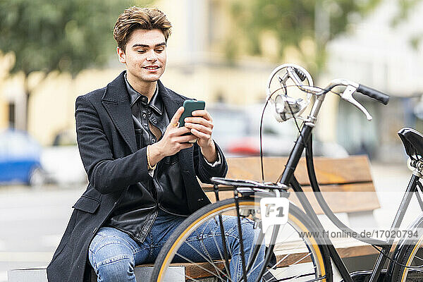 Junger Mann  der auf einer Bank in der Nähe eines Fahrrads in der Stadt sitzt und mit seinem Mobiltelefon SMS schreibt