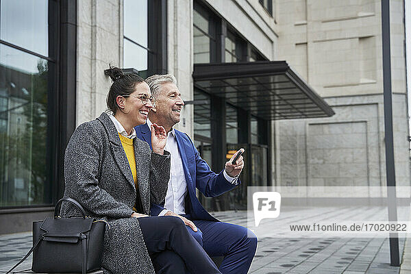 Fröhliche männliche und weibliche Unternehmer schauen weg  während sie mit ihrem Smartphone vor einem Gebäude sitzen