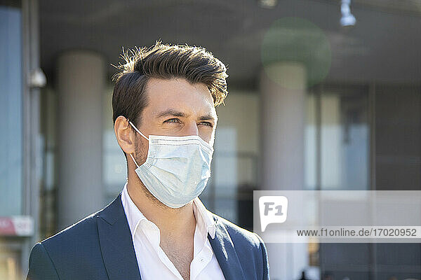 Nahaufnahme eines Geschäftsmannes mit Gesichtsmaske  der gegen ein Gebäude blickt