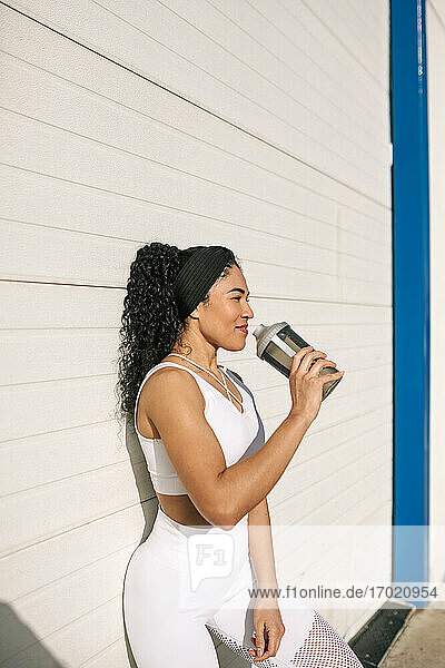 Lächelnde Sportlerin mit Wasserflasche an der Wand lehnend an einem sonnigen Tag