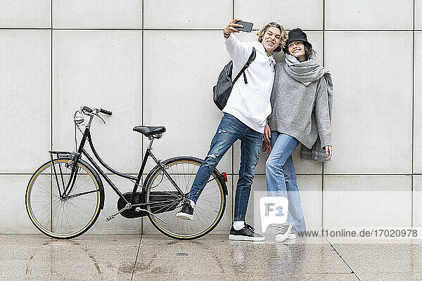 Mann  der ein Selfie macht  während seine Freundin mit dem Fahrrad auf dem Gehweg an der Wand steht