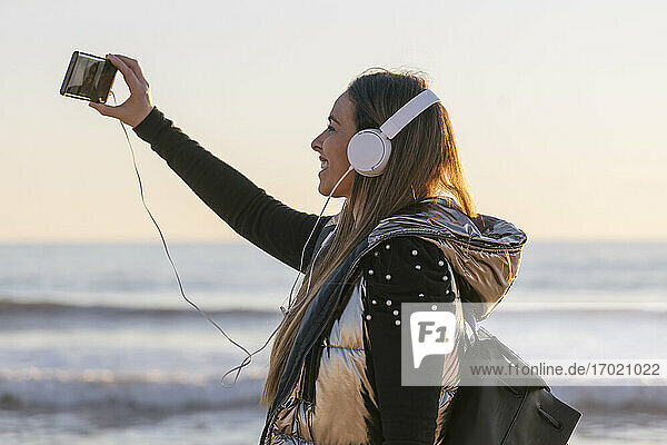 Woman taking selfie while listening music against sea during sunset