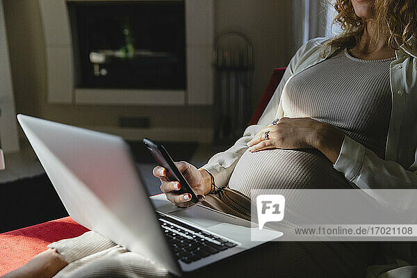 Schwangere Geschäftsfrau mit Mobiltelefon und Laptop auf dem Sofa zu Hause