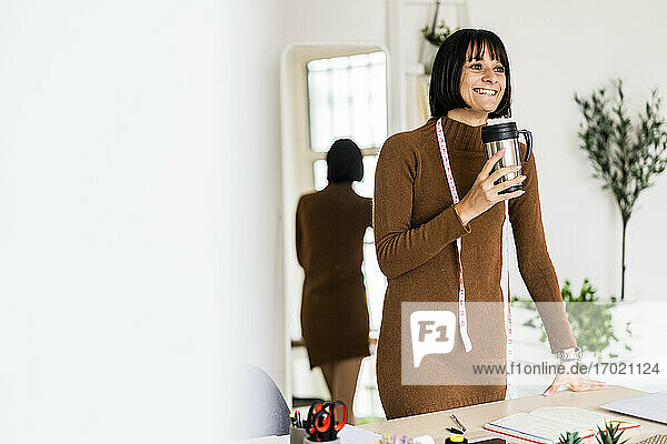 Fröhliche junge Stylistin träumt  während sie eine Kaffeetasse im Atelier hält