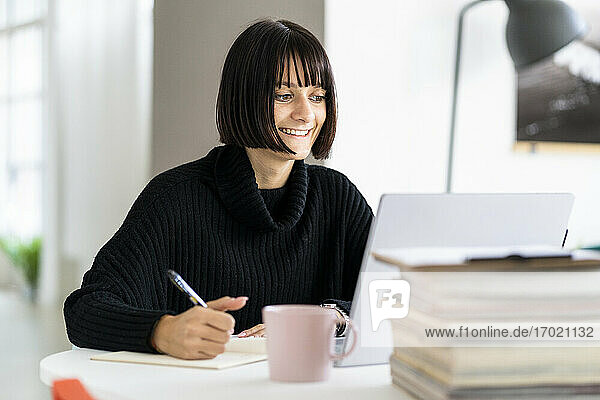 Smiling young female student with laptop writing while studying in study room