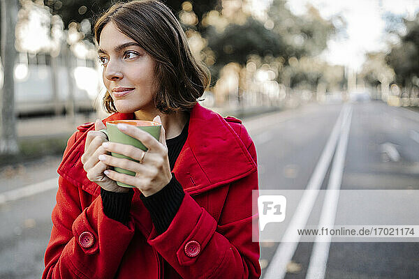 Nachdenkliche junge Frau in roter Jacke mit Kaffeetasse auf der Straße