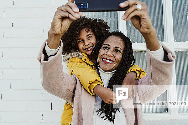 Mädchen  das seine Mutter umarmt und ein Selfie mit dem Smartphone an der Wand macht